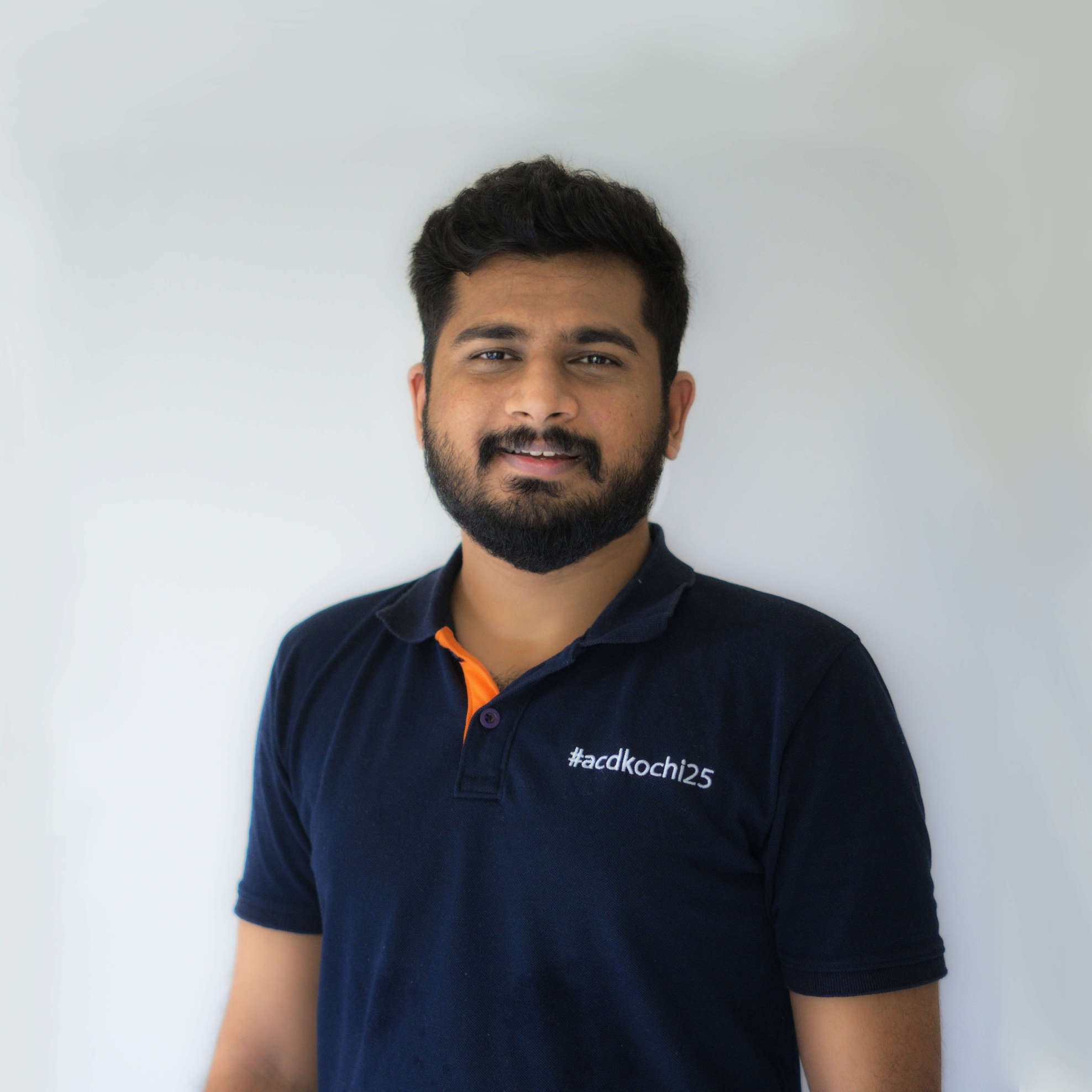 Photo of Vignesh in an off-white background. Vignesh is wearing a dark blue tshirt with an orange highlight on the strip where buttons are. The tshirt has #acdkochi embroidered on it on the chest towards the left.