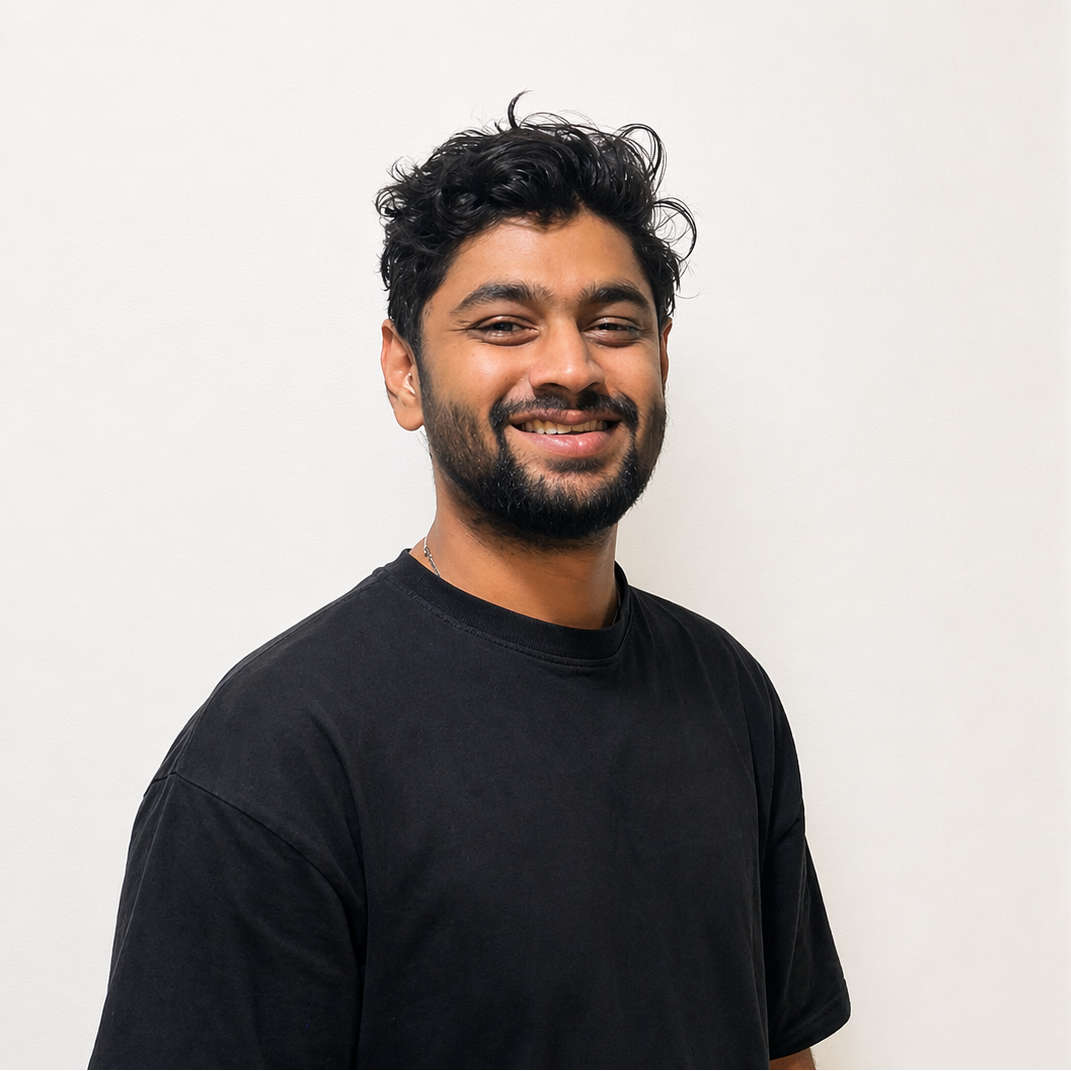 Photo of Vignesh in an off-white background. Vignesh is wearing a dark blue tshirt with an orange highlight on the strip where buttons are. The tshirt has #acdkochi embroidered on it on the chest towards the left.